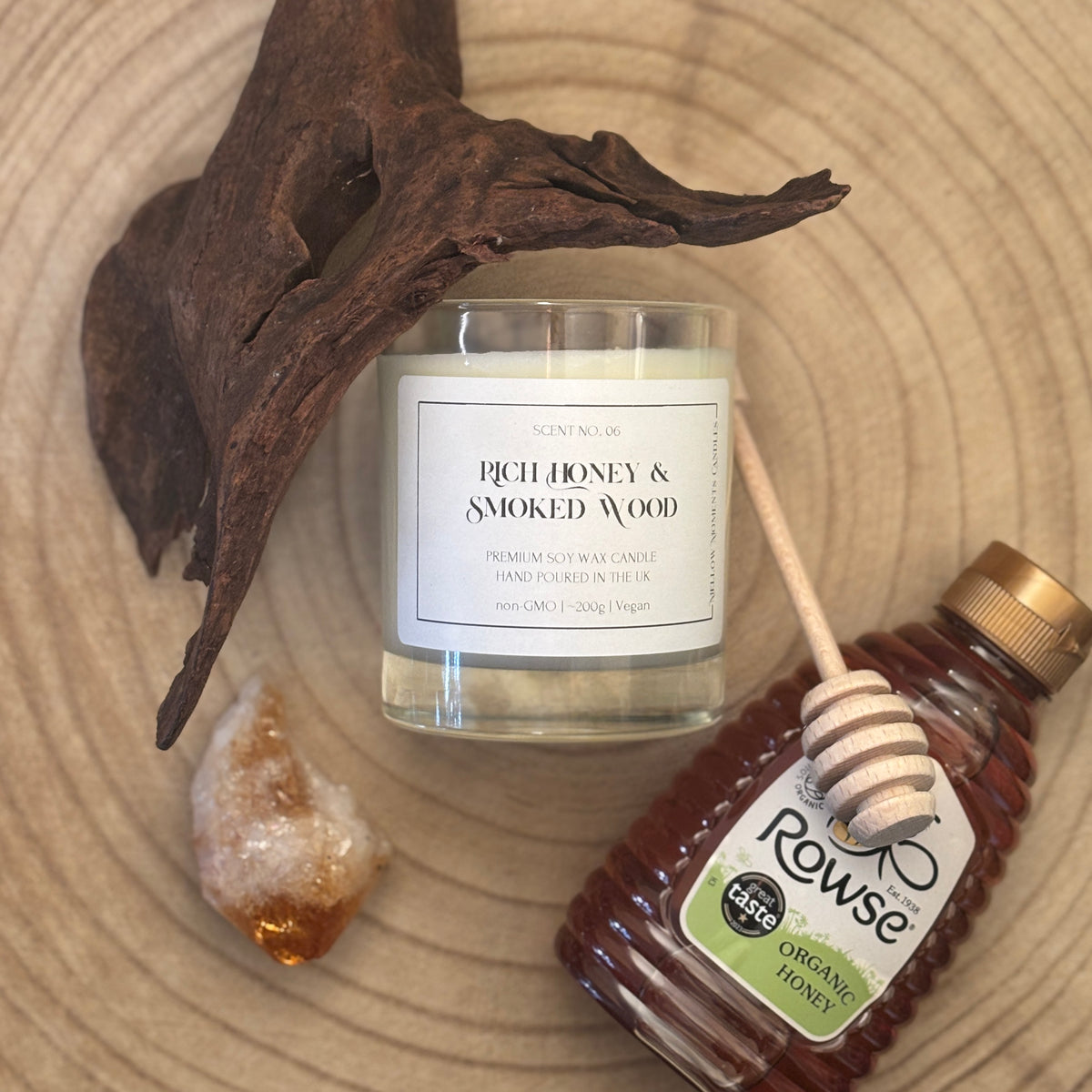Candle labelled 'Rich Honey & Smoked Wood' with a bottle of Rowse honey, honey dipper, amber crystal, and cedarwood on a wooden surface.