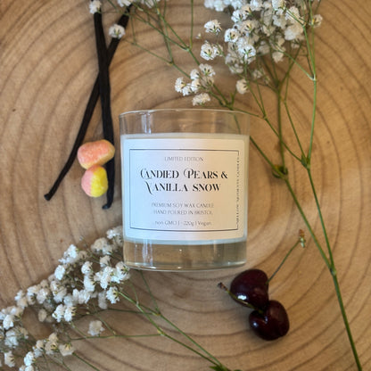 Candle labeled 'Candied Pears & Vanilla Snow' with vanilla beans, cherries, pear drops, and white flowers on a wooden surface
