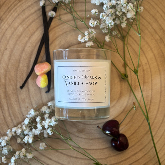 Candle labeled 'Candied Pears & Vanilla Snow' with vanilla beans, cherries, pear drops, and white flowers on a wooden surface