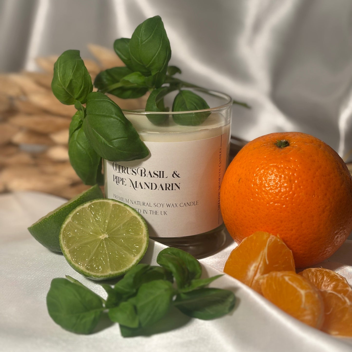 A handmade soy wax candle in a clear glass jar with a label reading 'Citrus Basil & Ripe Mandarin', accompanied by fresh basil leaves, a lime, an orange, and orange slices on a white silk surface.