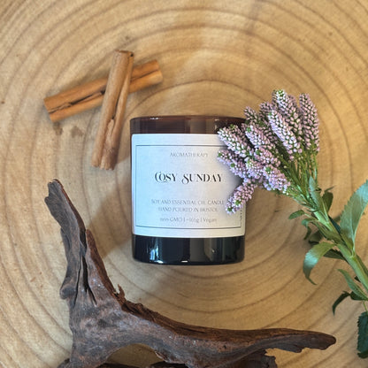 Candle labeled 'Cosy Sunday' on a wooden surface with cinnamon sticks, lavender and wood.