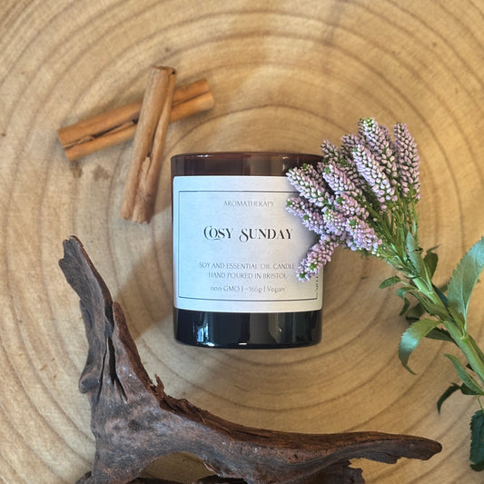 Candle labeled 'Cosy Sunday' on a wooden surface with cinnamon sticks, lavender and wood.