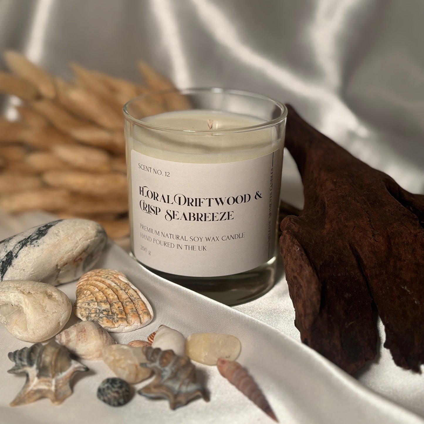 A handmade soy wax candle with the text 'FLORAL DRIFTWOOD & CRISP SEABREEZE' on it, surrounded by seashells and a piece of driftwood, with a background that includes a silk fabric and natural elements.