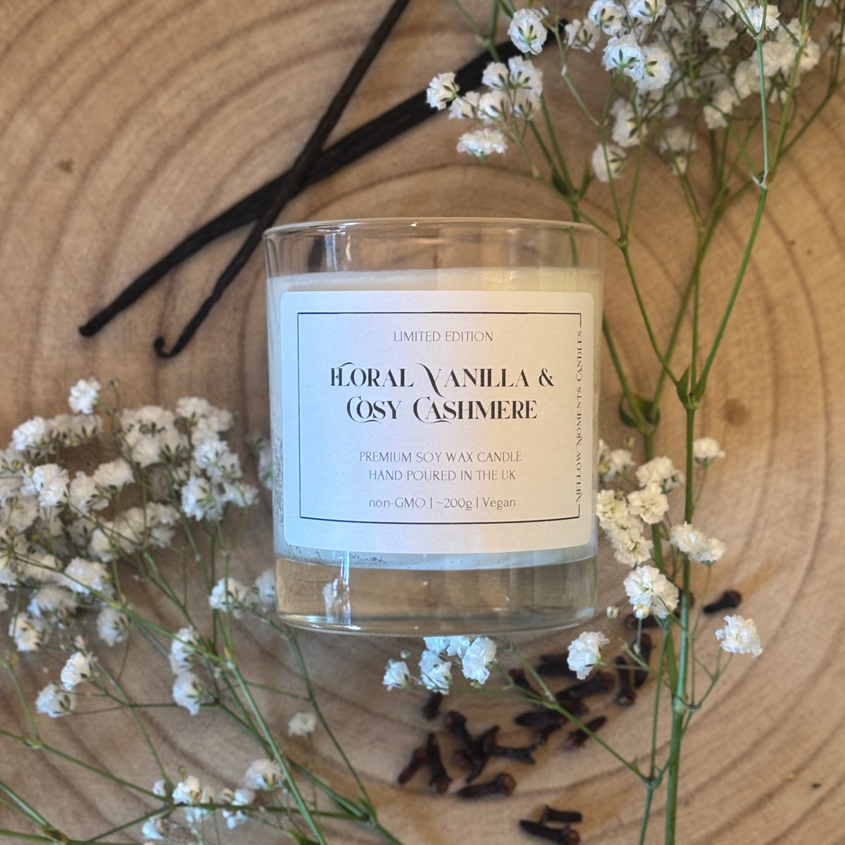 Candle labeled 'Floral Vanilla & Cosy Cashmere' surrounded by vanilla beans, cloves and small white flowers on a wooden surface.