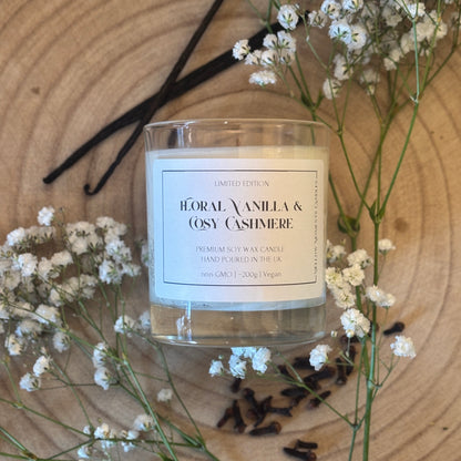 Candle labeled 'Floral Vanilla & Cosy Cashmere' surrounded by vanilla beans, cloves and small white flowers on a wooden surface.