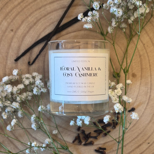 Candle labeled 'Floral Vanilla & Cosy Cashmere' surrounded by vanilla beans, cloves and small white flowers on a wooden surface.