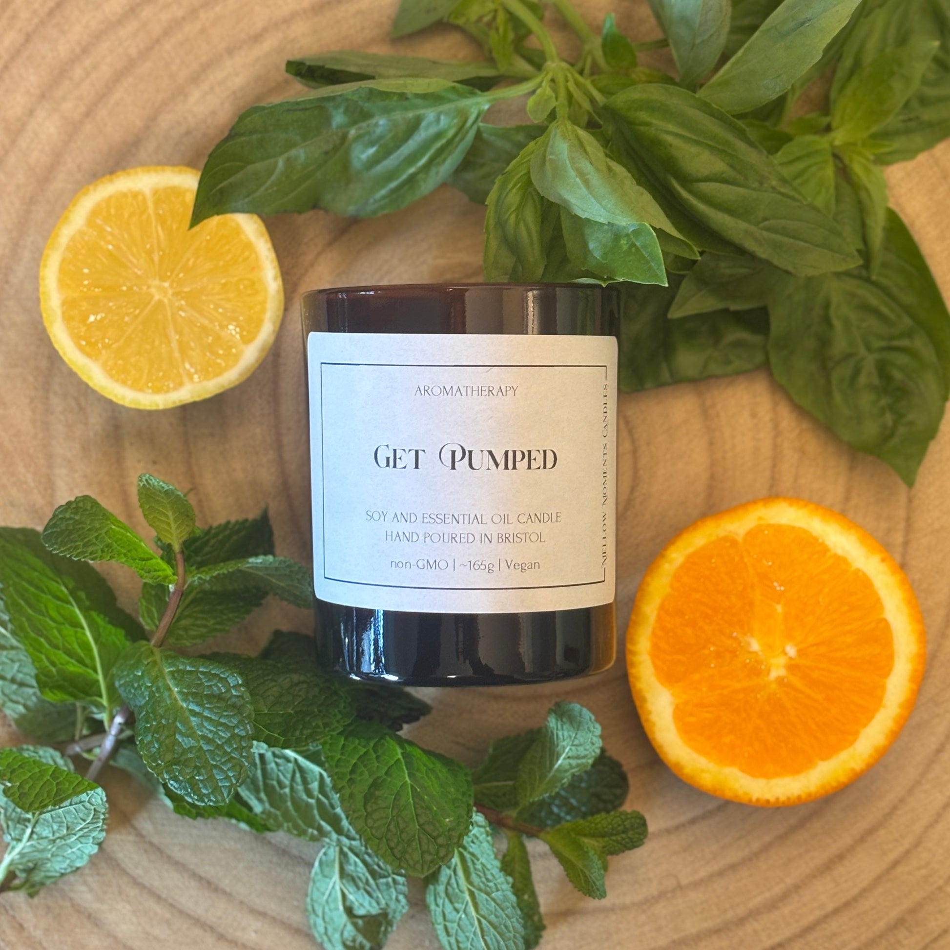Candle labeled 'GET PUMPED' surrounded by lemons, oranges, basil leaves, and mint leaves on a wooden surface