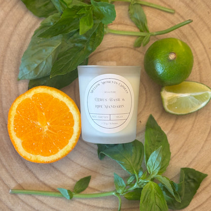 Frosted candle labelled "citrus basil and ripe mandarin" on a wooden surface with orange, lime, and basil leaves.