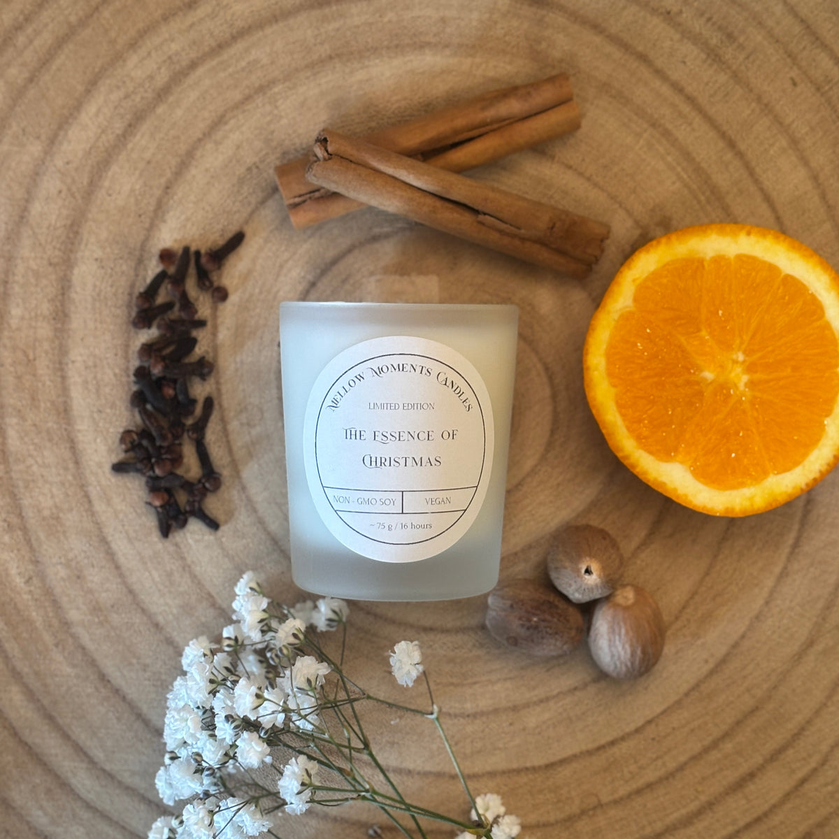 Frosted candle labeled 'The Essence of Christmas' surrounded by cinnamon sticks, cloves, an orange, nutmeg, and small flowers on a wooden surface.
