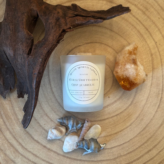 Frosted candle labelled " floral driftwood and crisp seabreeze" on a wooden surface with amber crystal, driftwood, and shells.
