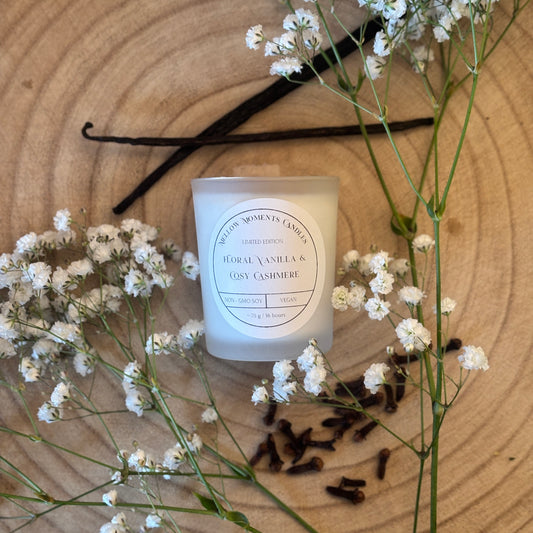 Frosted candle labelled "floral vanilla & cosy cashmere" with vanilla bean, cloves, and white flowers on a wooden surface 