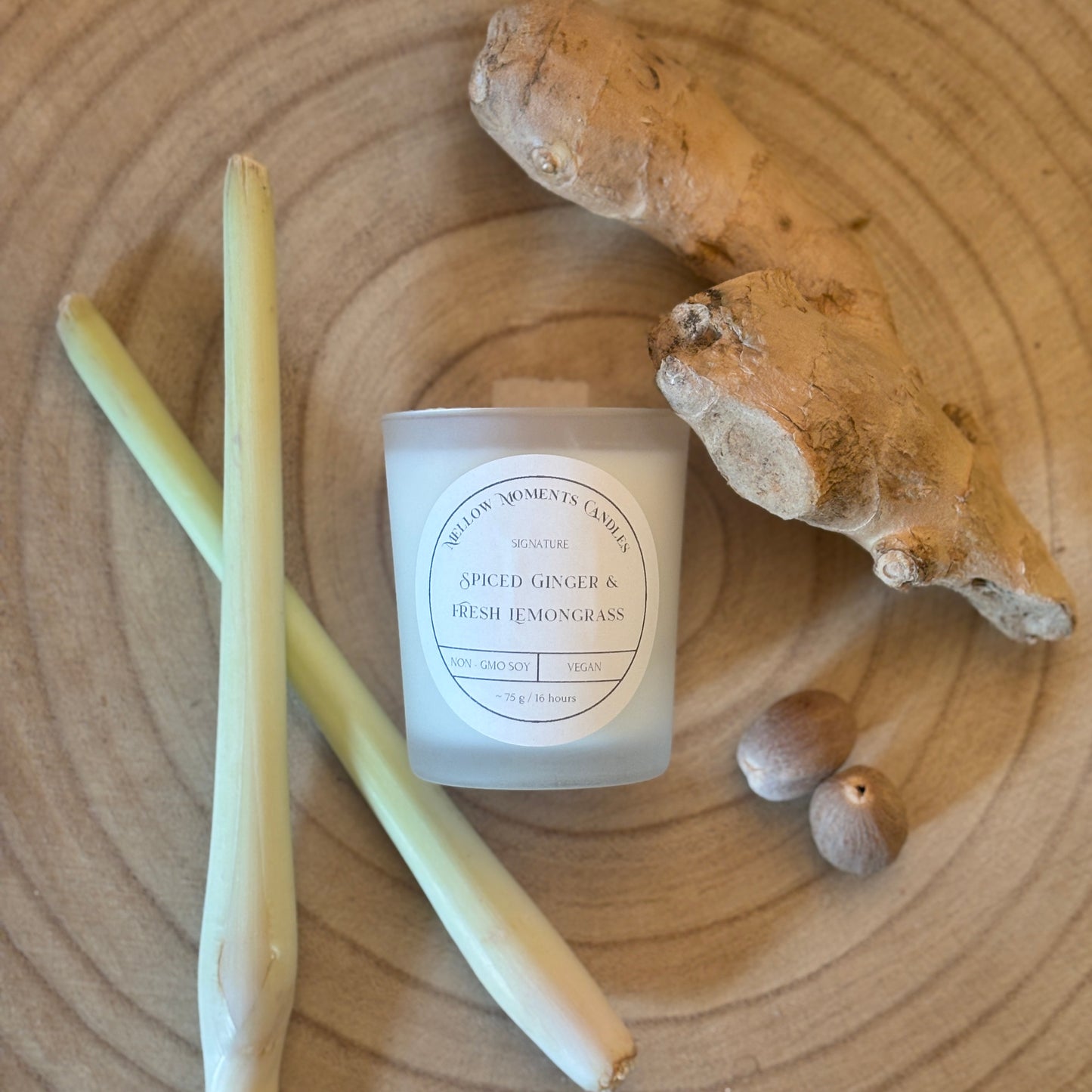 Candle labelled 'Spiced Ginger & Fresh Lemongrass' with ginger root, nutmeg, and lemongrass stalk on a wooden surface