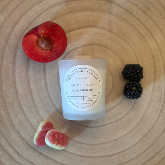 Frosted candle labelled 'Sweet Plum & Wild Rhubarb' on a wooden surface with a plum, Rhubarb and cream sweets, and blackberries.