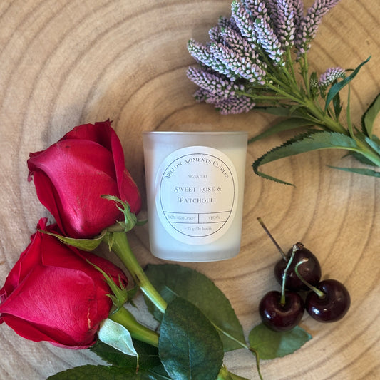 Frosted candle labelled with "Sweet rose & Patchouli" with red roses, lavender, and cherries on a wooden surface