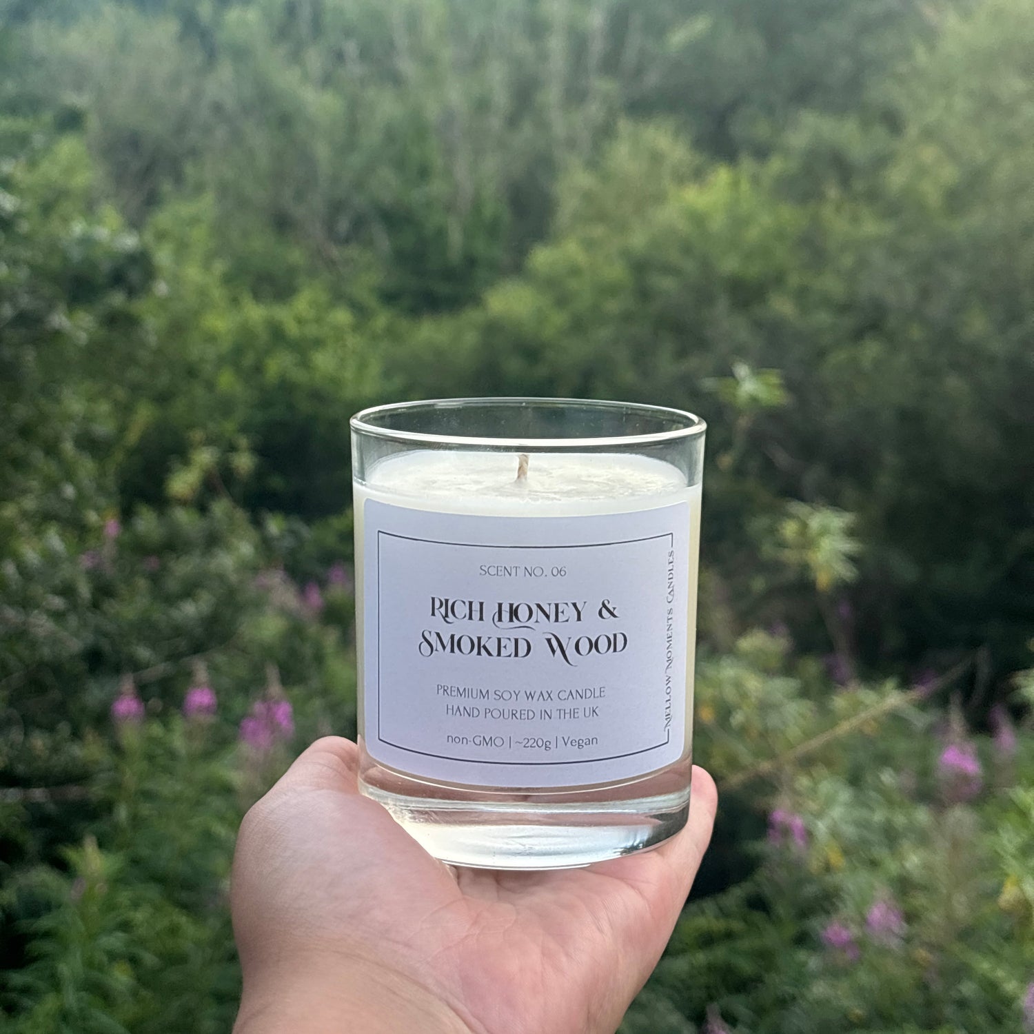 Candle labeled 'Rich Honey & Smoked Wood' held in front of a forest background