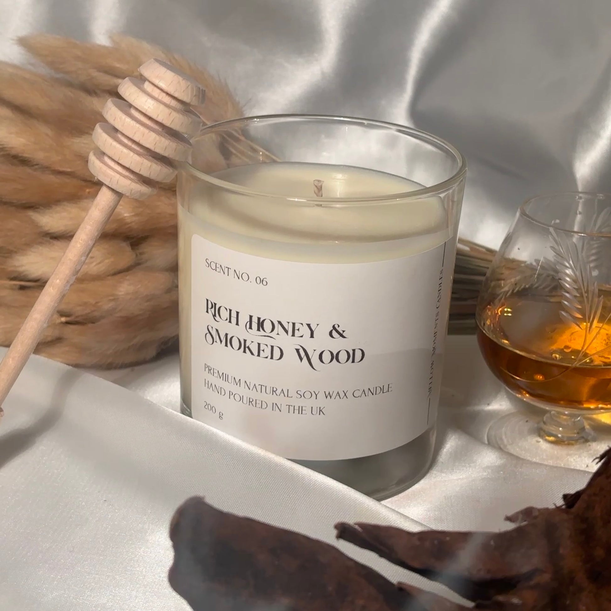 A clear glass soy wax candle with a label reading 'Rich Honey & Smoked Wood', accompanied by a honey dipper and a glass of cognac, set on a white silk fabric surface and a piece of driftwood placed in front.