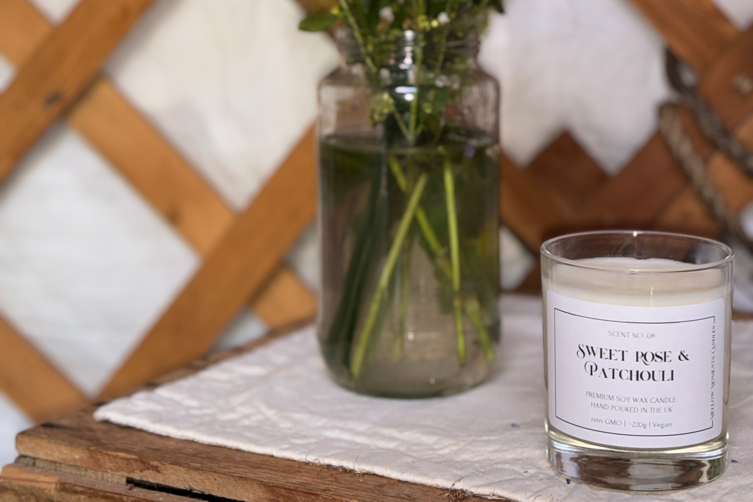 Vase of flowers and a candle scented 'Sweet Rose & Patchouli' on a wooden surface with a wooden lattice background.