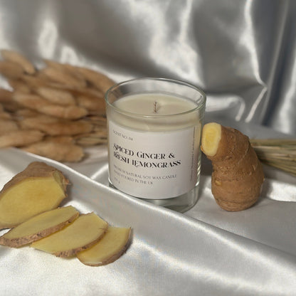 A clear glass soy wax candle with a label reading 'Spiced Ginger & Fresh Lemongrass', surrounded by slices of ginger and a piece of lemongrass, all placed on a white satin fabric surface.