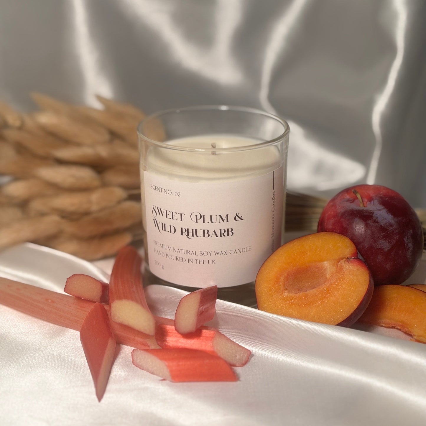 A glass candle with white soy wax written as 'Sweet Plum & Wild Rhubarb', surrounded by a piece of rhubarb and a plum on a white silk surface.