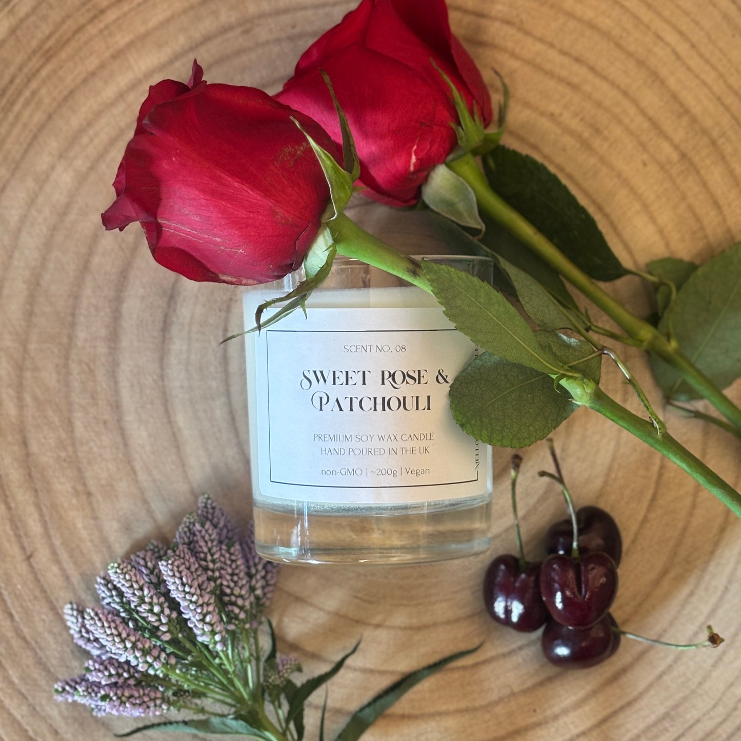 Candle labeled 'Sweet Rose & Patchouli' with red roses, cherries, and lavender on a wooden surface
