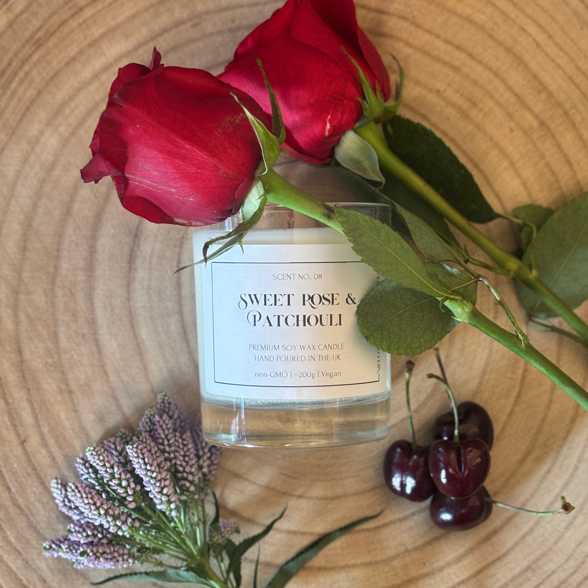 Candle labeled 'Sweet Rose & Patchouli' with red roses, cherries, and lavender on a wooden surface