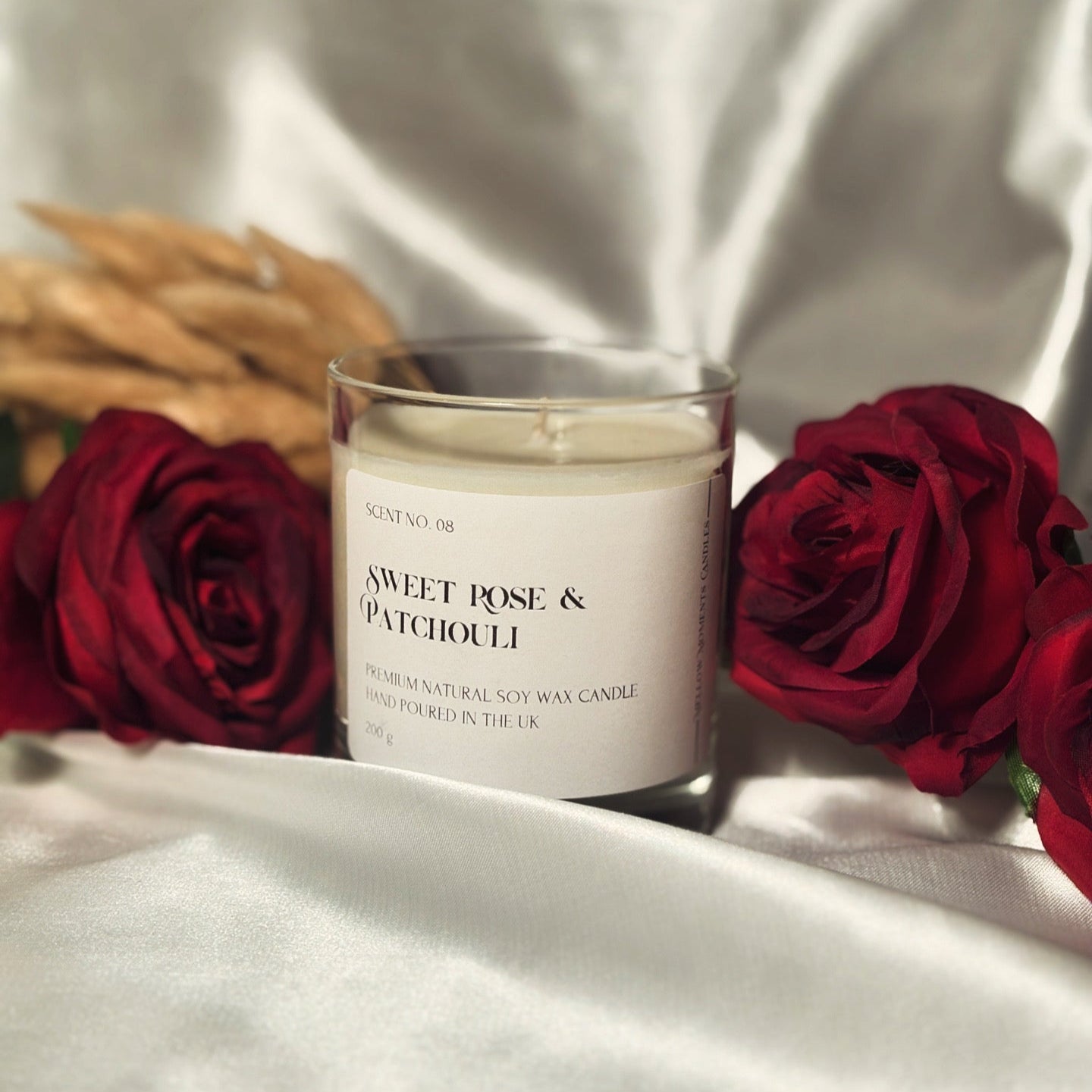 A handmade soy wax candle in a clear glass candle jar with a white label, placed on a white silk surface with three red roses and some dried flowers in the background.