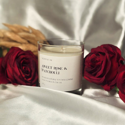 A handmade soy wax candle in a clear glass candle jar with a white label, placed on a white silk surface with three red roses and some dried flowers in the background.