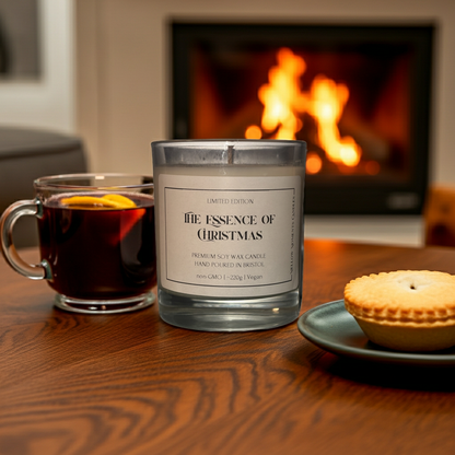 Candle labeled 'The Essence of Christmas' with a mug of mulled wine and a minced pie on a wooden table in front of a fireplace.