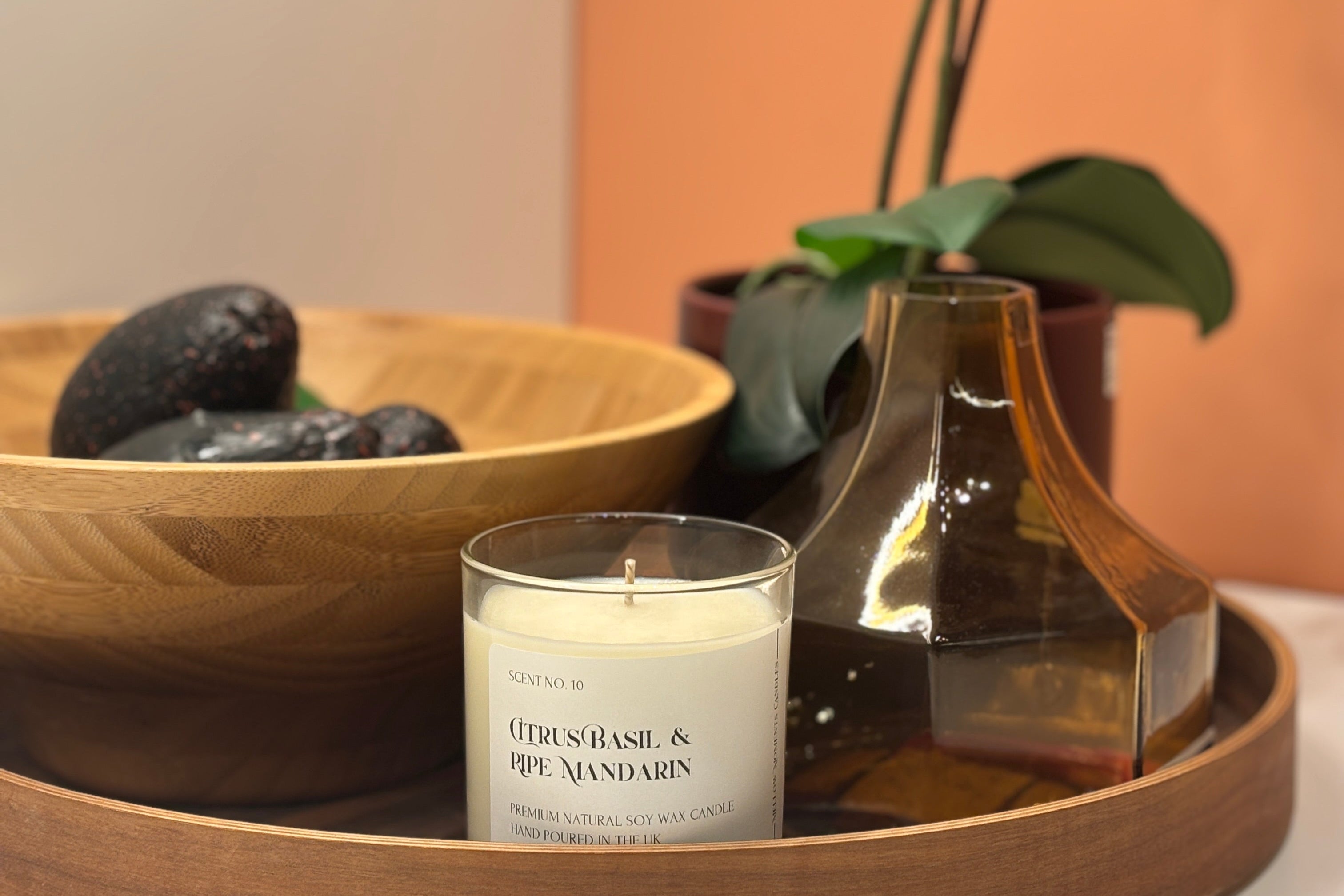 A basil, lime and mandarin scented candle on a wooden tray with a bowl filled with avocado, a flask, and a vase with an orchid in it in the background.