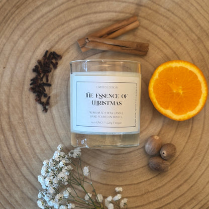 Candle labeled 'The Essence of Christmas' on a wooden surface with cinnamon sticks, an orange, nutmeg, clove, and flowers.
