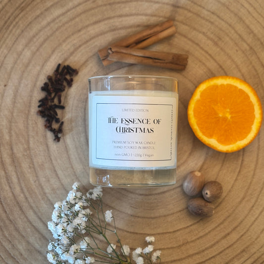 Candle labeled 'The Essence of Christmas' on a wooden surface with cinnamon sticks, an orange, nutmeg, clove, and flowers.