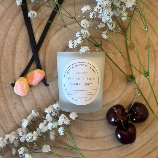 Mini candle labeled 'Candied Pears & Vanilla Snow' with vanilla beans, cherries, pear drops, and flowers on a wooden surface
