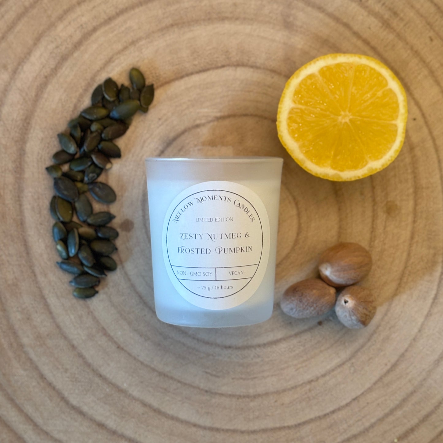 Frosted candle labeled 'Zesty Nutmeg & Frosted Pumpkin' on a wooden surface with pumpkin seeds, a lemon, and nutmeg.