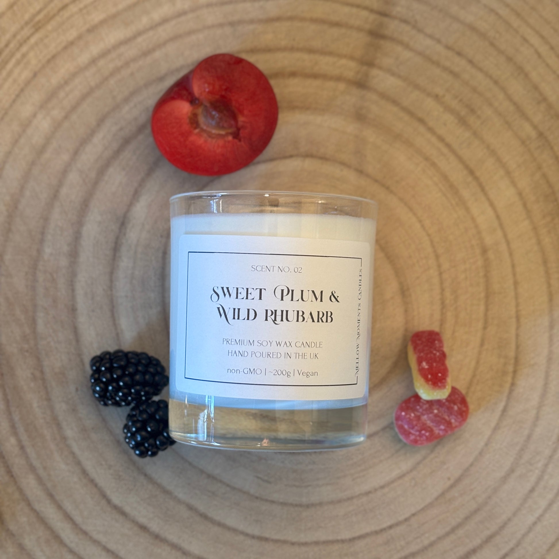Candle labeled 'Sweet Plum & Wild Rhubarb' with a plum, blackberry, and rhubarb cream sweets on a wooden surface