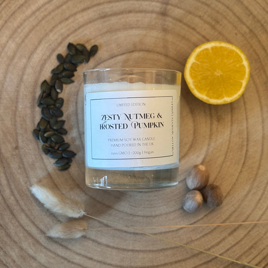 Candle labeled 'Zesty Nutmeg & Frosted Pumpkin' on a wooden surface with pumpkin seeds, a lemon, and nutmeg.